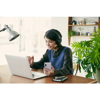 Eine lächelnde Person mit Kopfhörern sitzt an einem Tisch, nutzt einen Laptop und hält ein Smartphone. Im Hintergrund sind Pflanzen sichtbar.