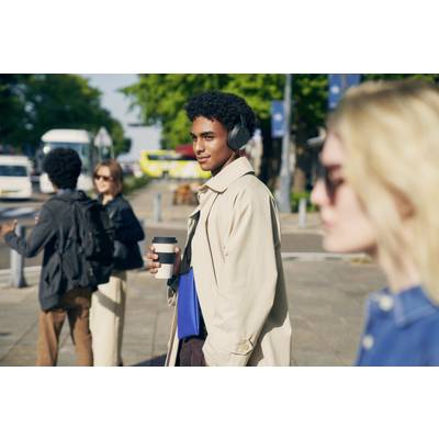 Eine Person mit Kopfhörern geht eine belebte Straße entlang, hält einen Kaffee und trägt eine Mappe. Andere Menschen sind unscharf im Hintergrund.