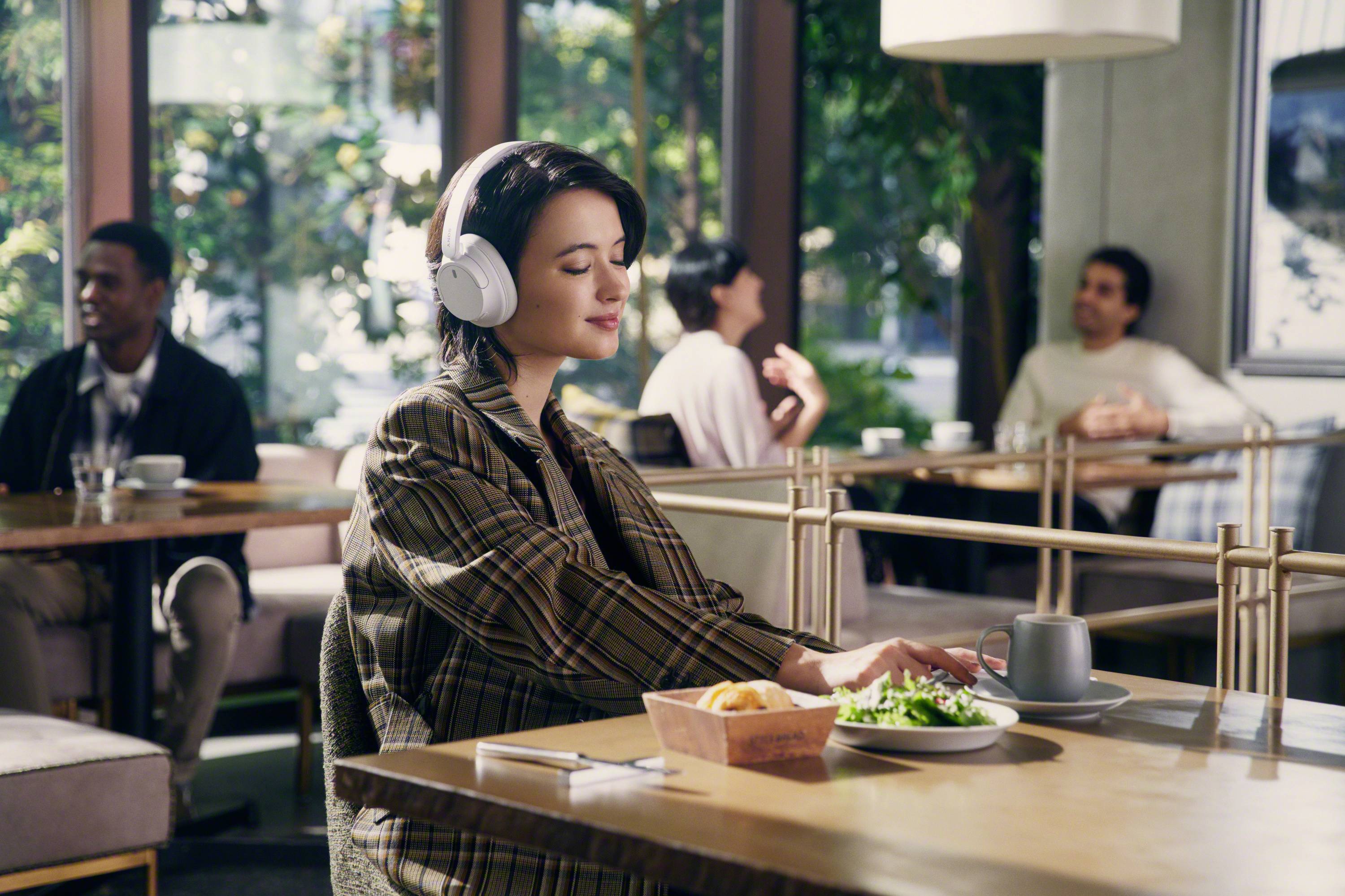 Eine Person sitzt in einem Café, trägt Kopfhörer und genießt eine Mahlzeit auf einem Tisch. Andere Gäste unterhalten sich im Hintergrund.