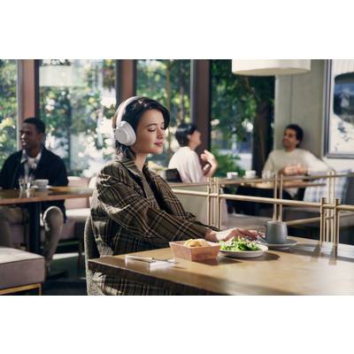 Eine Person sitzt in einem Café, trägt Kopfhörer und genießt eine Mahlzeit auf einem Tisch. Andere Gäste unterhalten sich im Hintergrund.