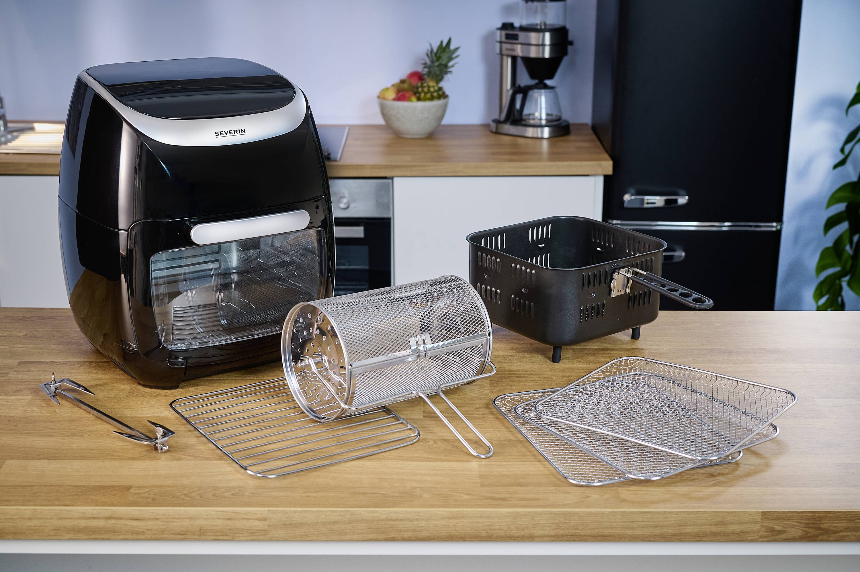 Heißluftfritteuse mit Zubehör in einer Küche. Zubehörteile umfassen Gitter, Korb und Zange. Küche mit modernen Geräten im Hintergrund.