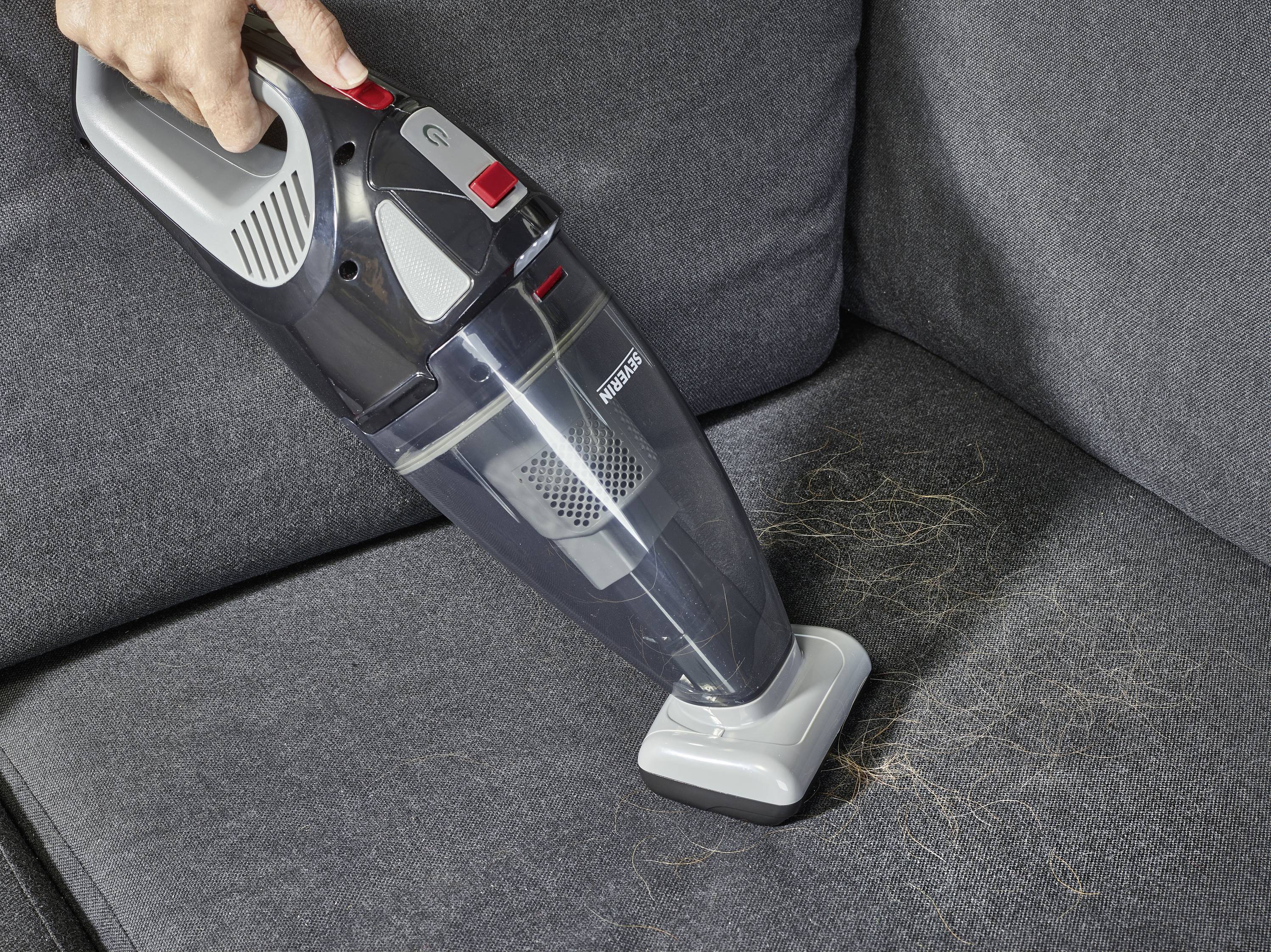 Eine Person saugt Haare von einem grauen Sofa mit einem kabellosen Handstaubsauger ab.