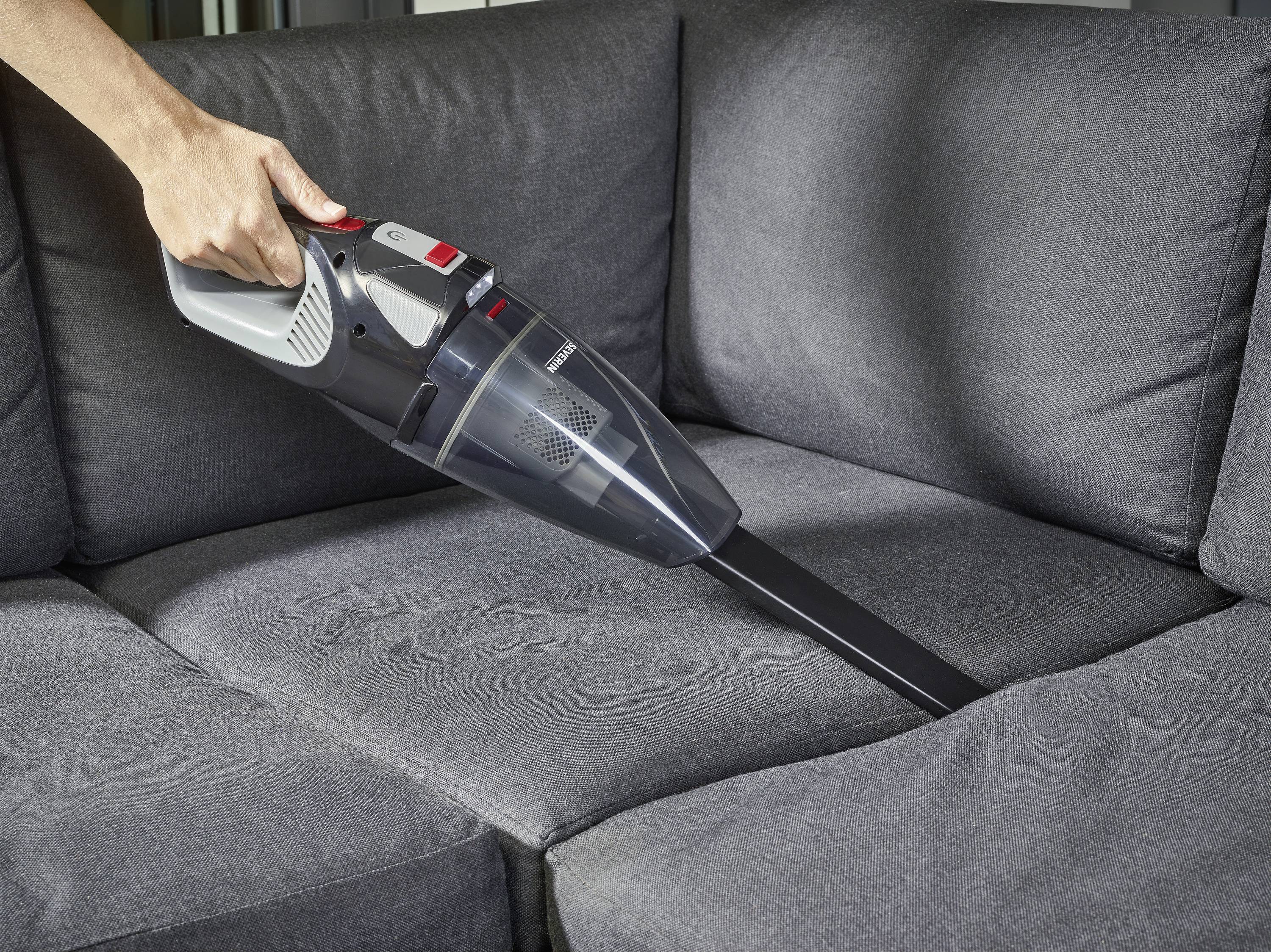 Eine Hand führt einen Handstaubsauger über ein graues Sofa, um es zu reinigen.