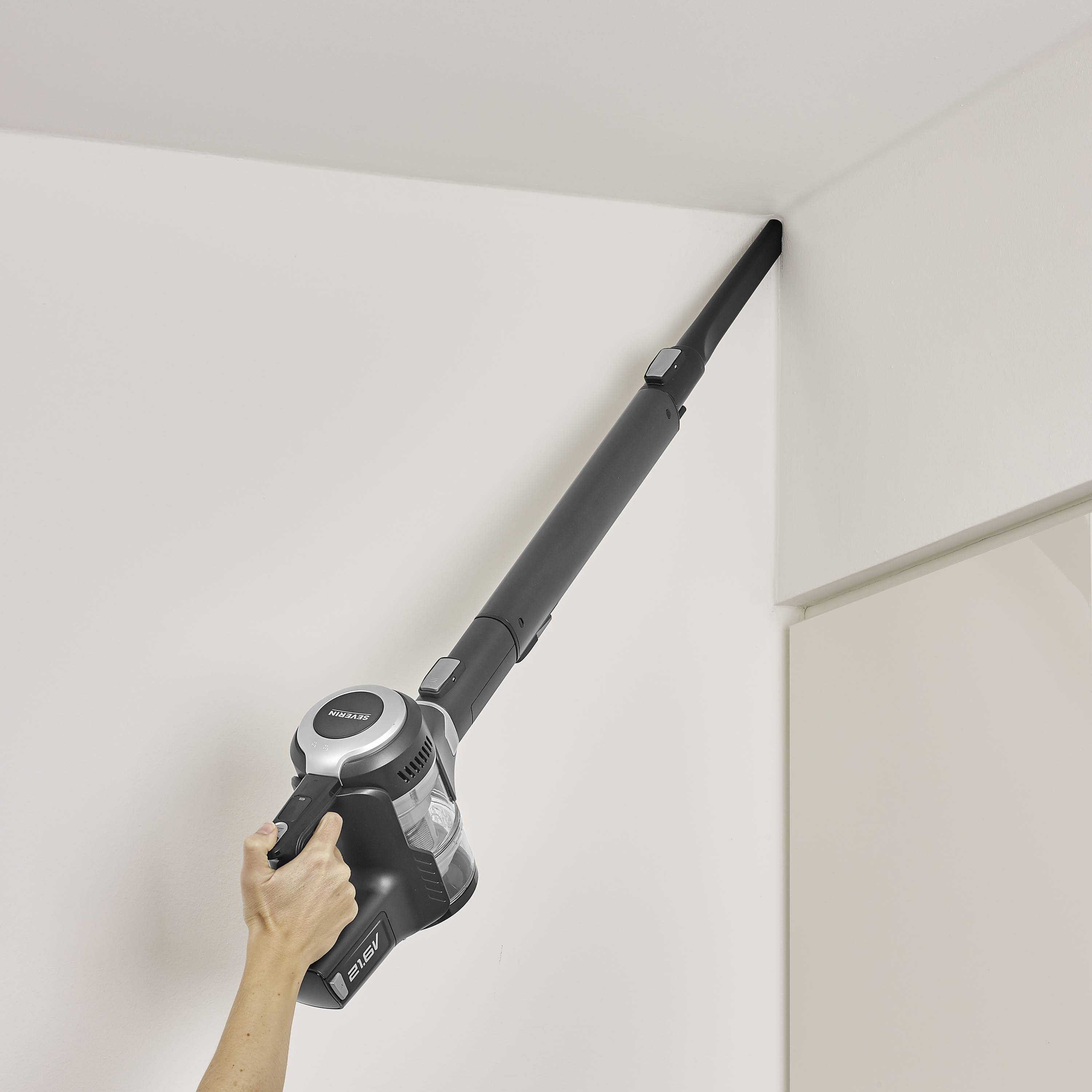 Eine Hand hält einen Stabstaubsauger, der eine Ecke an der Zimmerdecke saugt. Der Fokus liegt auf der Reinigung des schwer erreichbaren Bereichs.