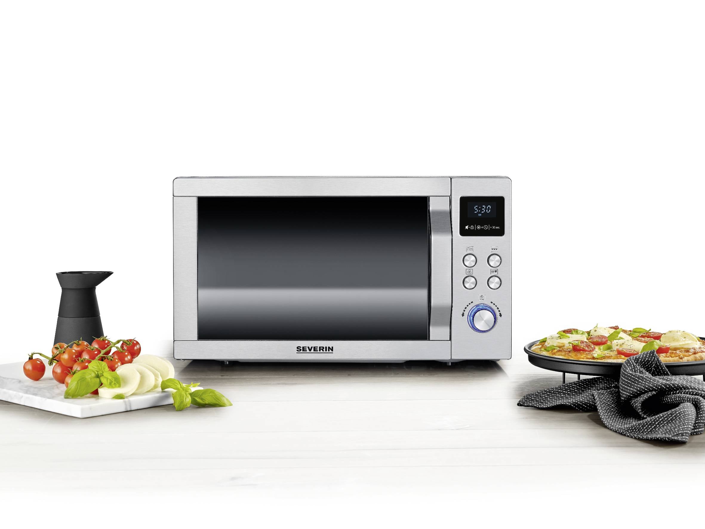 Silberne Mikrowelle auf Küchentisch, umgeben von gestapelten Tomaten, Zwiebeln, Basilikum und einer Pizza auf einem Teller.