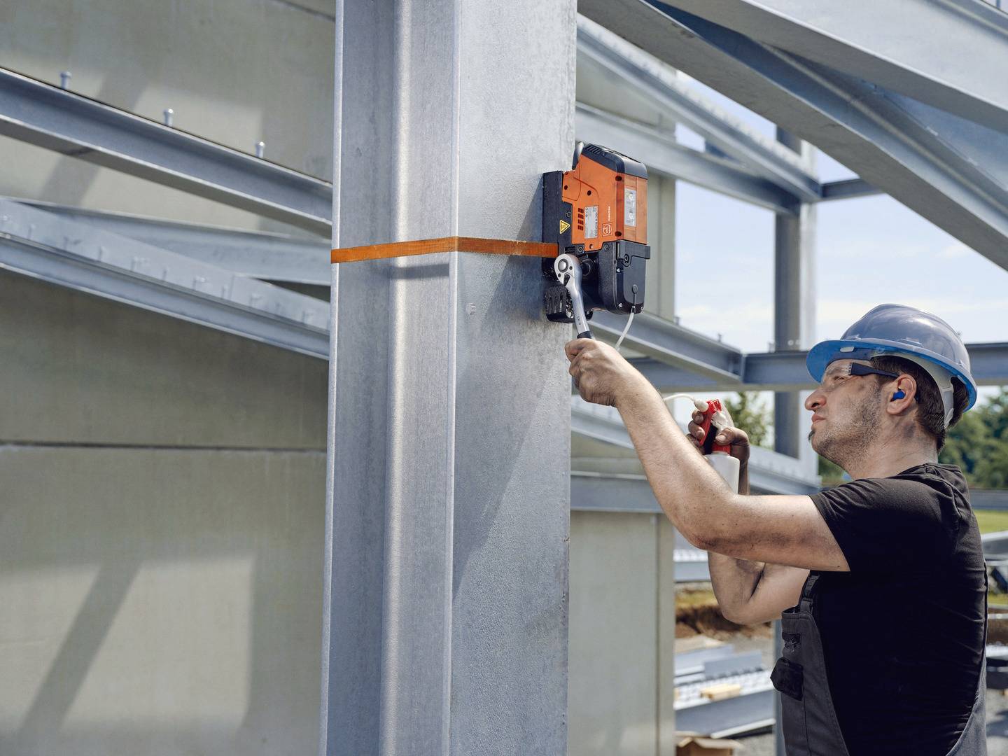 Ein Bauarbeiter mit Helm befestigt mit einem Schraubenschlüssel ein Stück Stahl an einer großen Stahlstütze einer Baustelle.