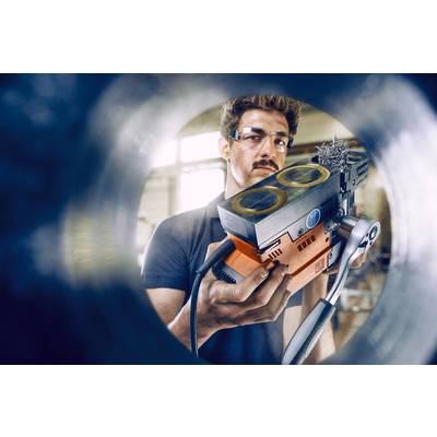 Ein Mann mit Schutzbrille hält ein orange-schwarzes Gerät, möglicherweise ein technisches Werkzeug. Er befindet sich in einer industriellen Umgebung.