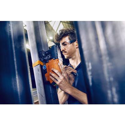 Ein Mann mit Schutzbrille überprüft mit einem Messgerät die Struktur eines Metallträgers in einem industriellen Umfeld.