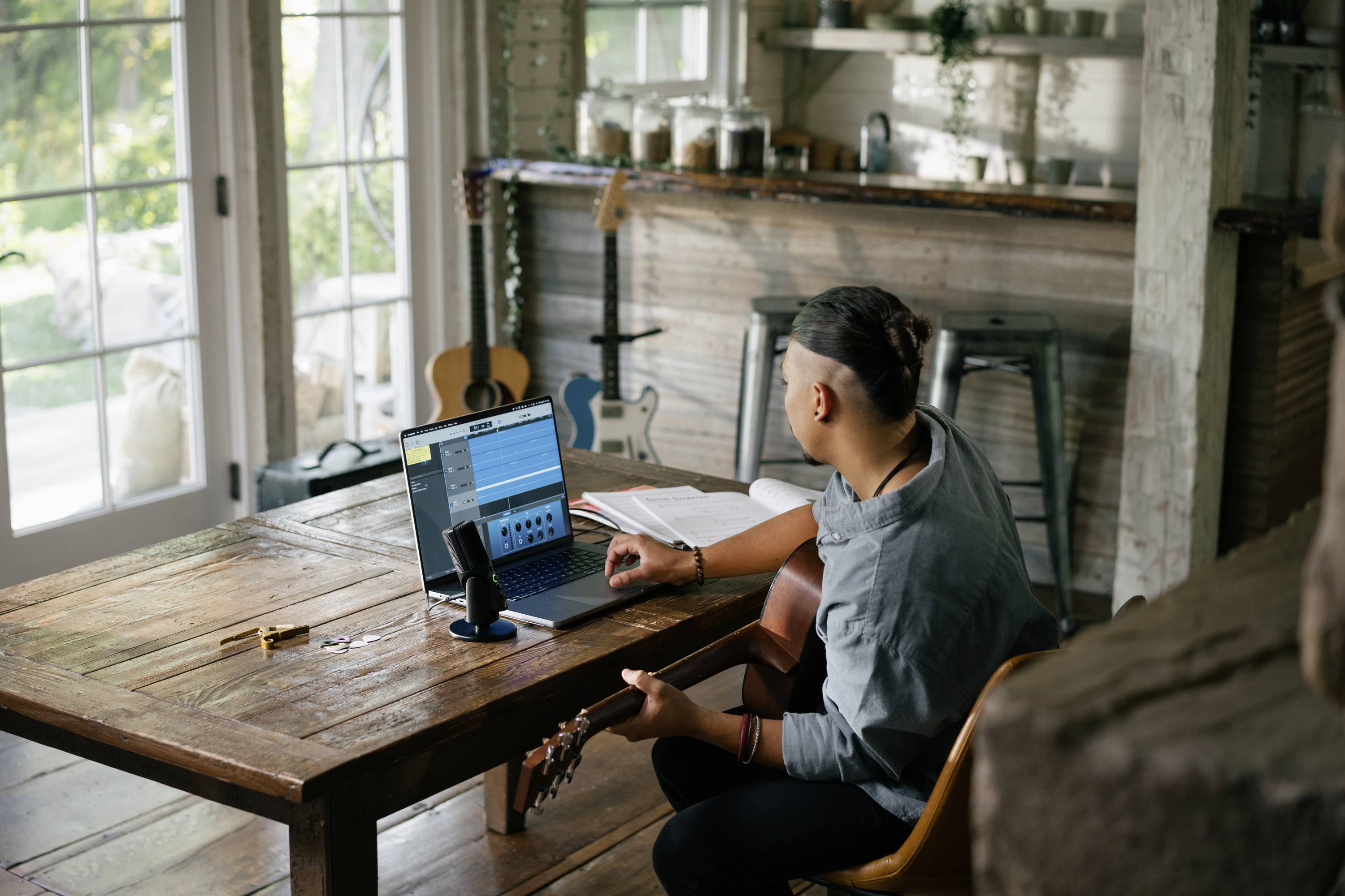 Mensch sitzt an einem Holztisch mit Laptop und Mikrofon, spielt Gitarre. Fenster im Hintergrund zeigt sonnigen Garten. Kreativer Arbeitsbereich.