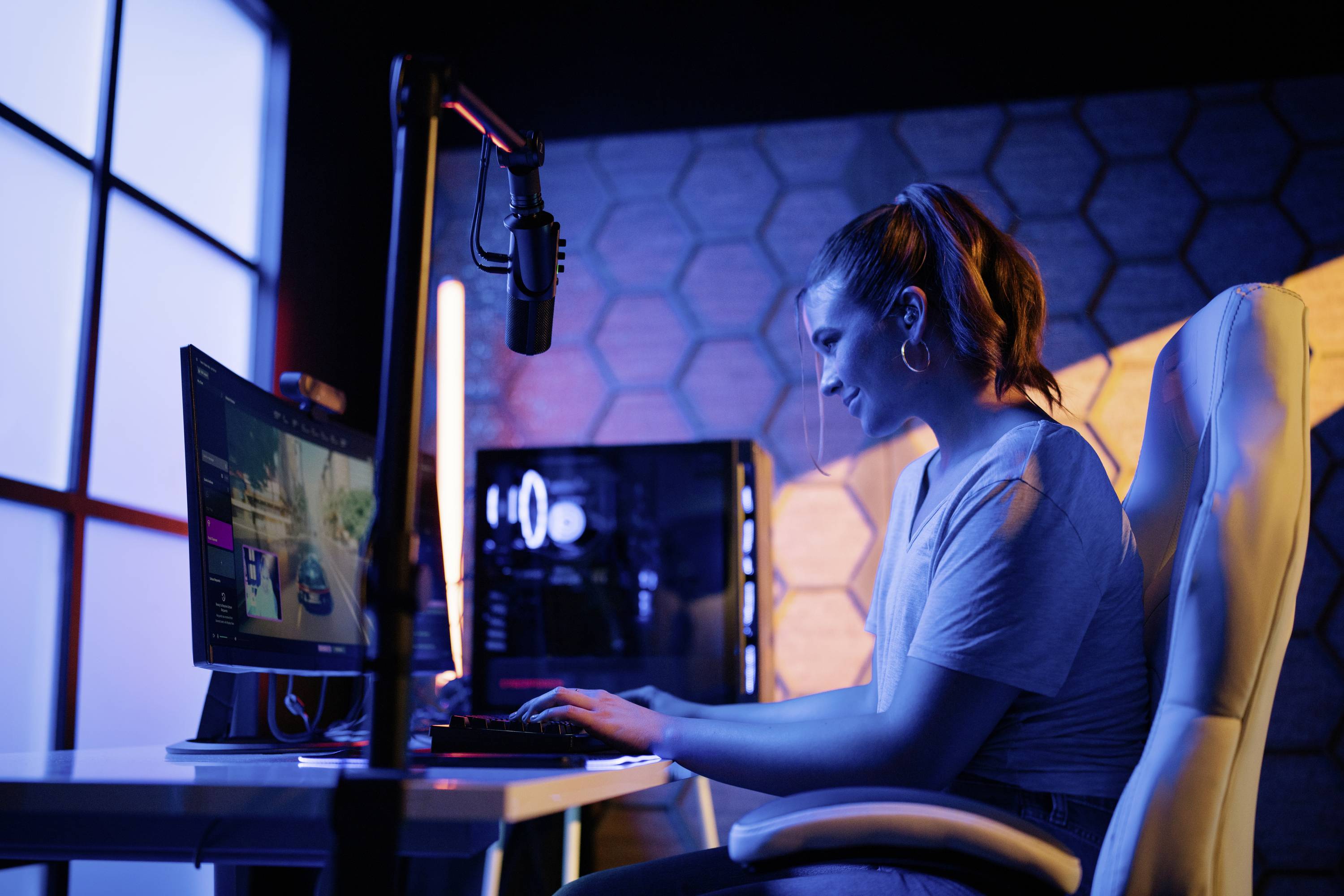 Eine Frau sitzt an einem Schreibtisch und spielt ein Videospiel auf einem Computer. Ein Mikrofon hängt vor ihr. Die Umgebung ist bunt beleuchtet.
