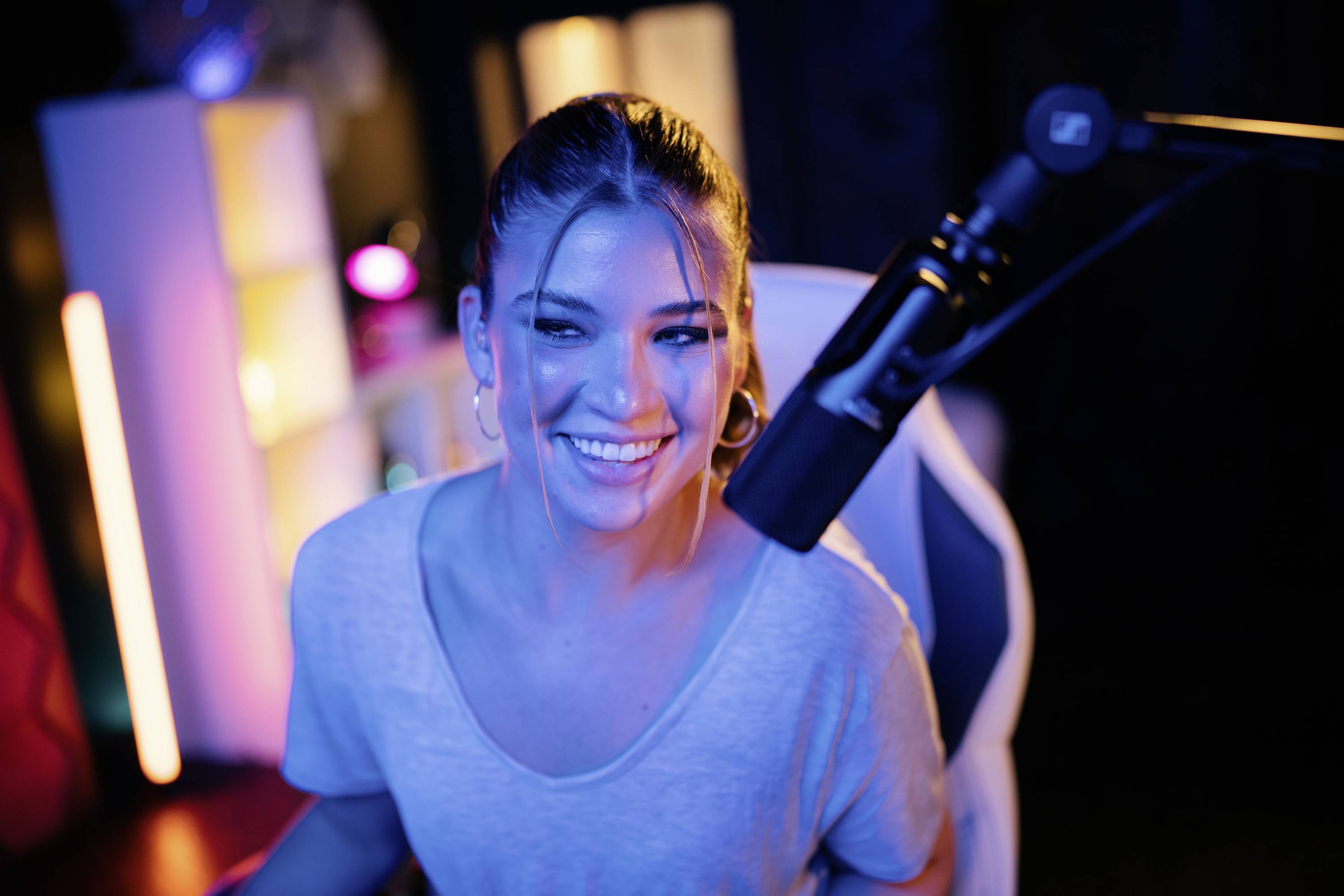 Eine lächelnde Frau sitzt vor einem Mikrofon in einem bunt beleuchteten Raum, vermutlich ein Streaming- oder Aufnahmestudio.
