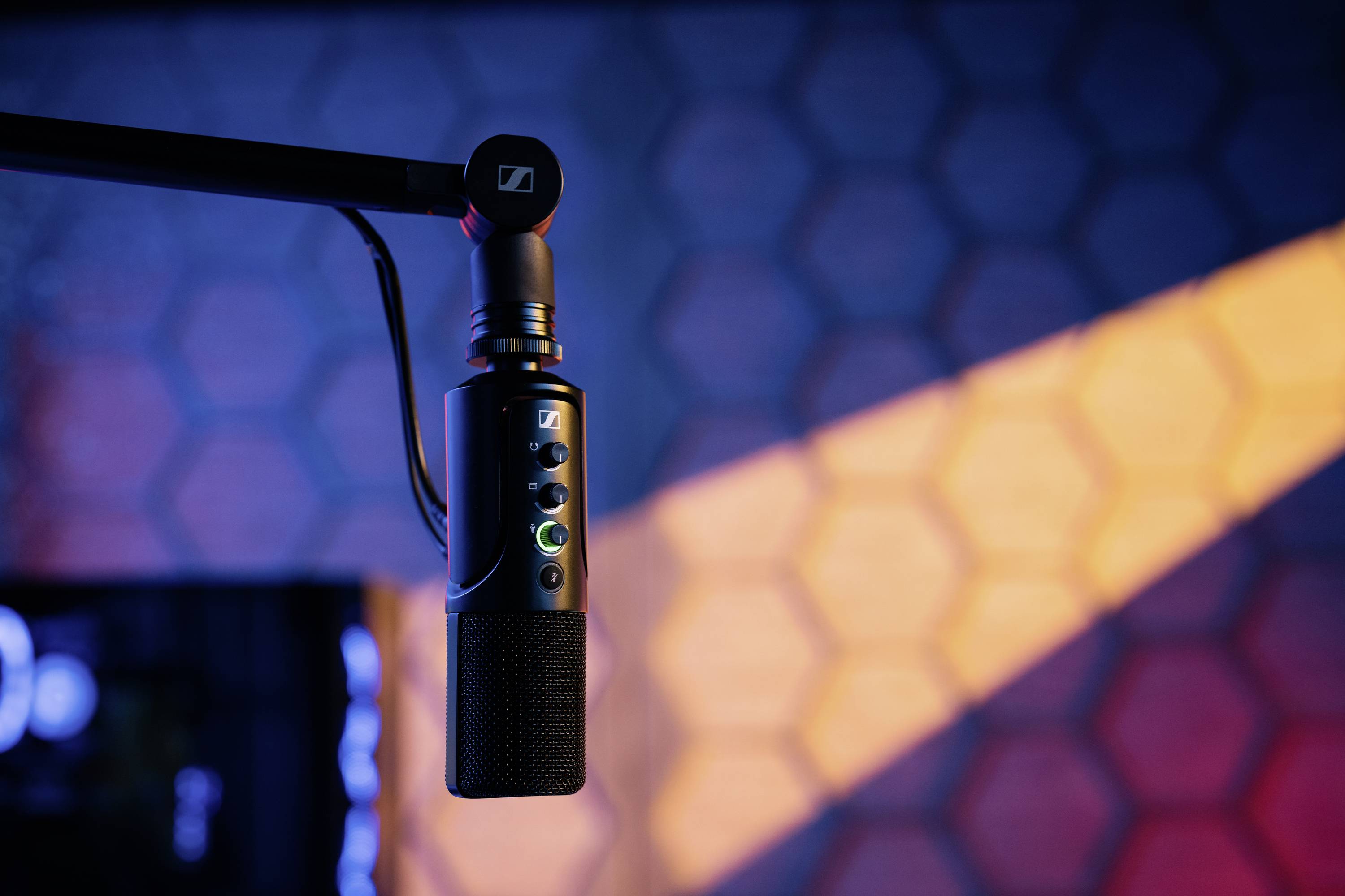 Ein Mikrofon hängt an einem Arm vor einer Wand mit Wabenmuster. Es wird von buntem Licht in Blau- und Orangetönen beleuchtet.