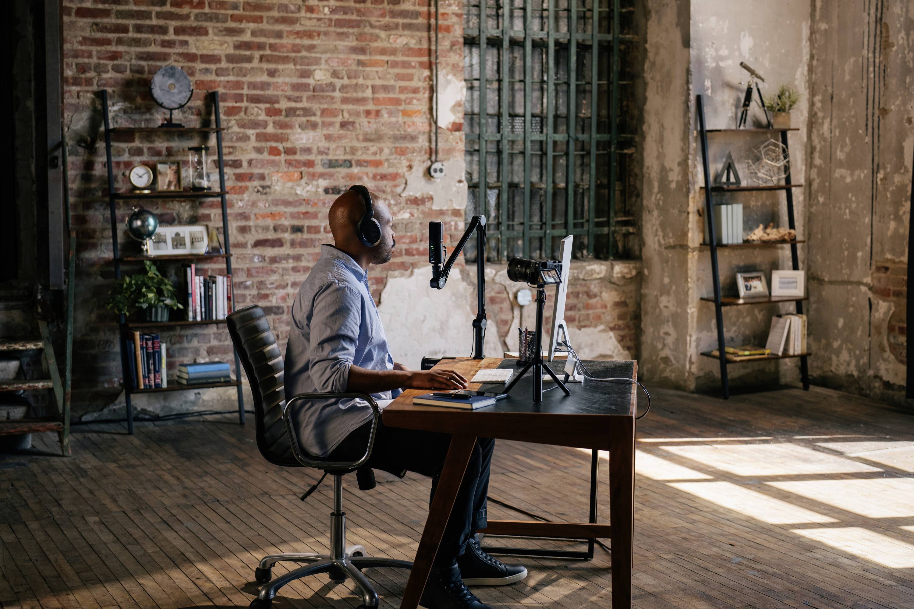 Eine Person sitzt an einem Schreibtisch in einem industriell gestalteten Raum, arbeitet am Computer und trägt Kopfhörer. Holzfußboden und Ziegelwände.