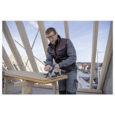 Eine Person in Schutzausrüstung bedient eine Kreissäge an einem Stück Holz auf einer Baustelle mit einer Stadt im Hintergrund.
