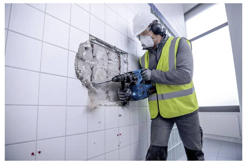 Ein Bauarbeiter in Schutzausrüstung verwendet eine Bohrmaschine, um Fliesen von einer Wand zu entfernen und damit eine Renovierungsaktivität zu veranschaulichen.