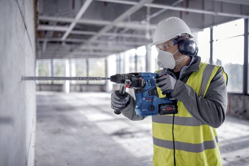 Ein Bauarbeiter mit Sicherheitsausrüstung bohrt in eine Betonwand in einem teilweise fertiggestellten Gebäude, umgeben von Metallträgern.