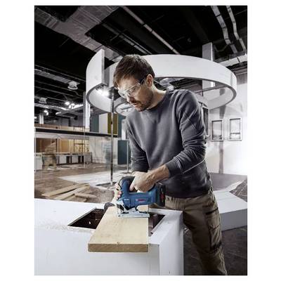 Ein Mann mit Sicherheitsbrille bedient eine Kreissäge und schneidet ein Holzbrett in einer industriellen Werkstattumgebung.