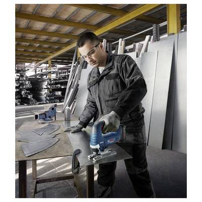 Ein Arbeiter in Schutzausrüstung benutzt eine Stichsäge zum Schneiden eines Metallblechs in einer Fabrik mit Stapeln von Metall im Hintergrund.