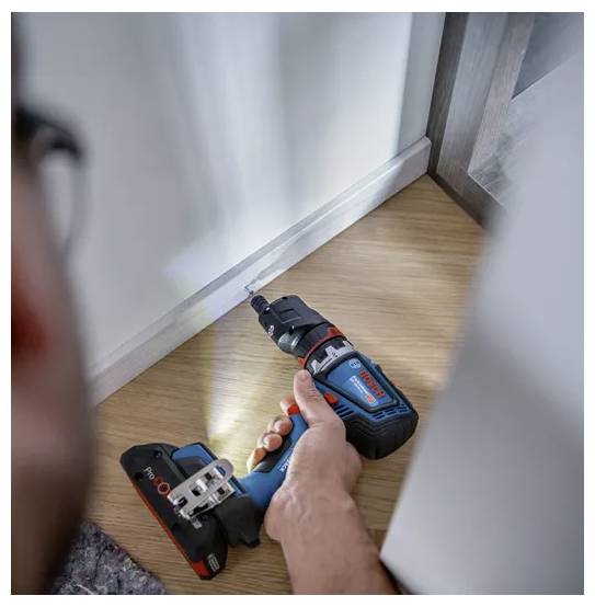 Eine Person, die eine kabellose Bohrmaschine verwendet, um eine hellgefärbte Sockelleiste an der Ecke eines Holzbodens in der Nähe der unteren Wandkante zu befestigen.