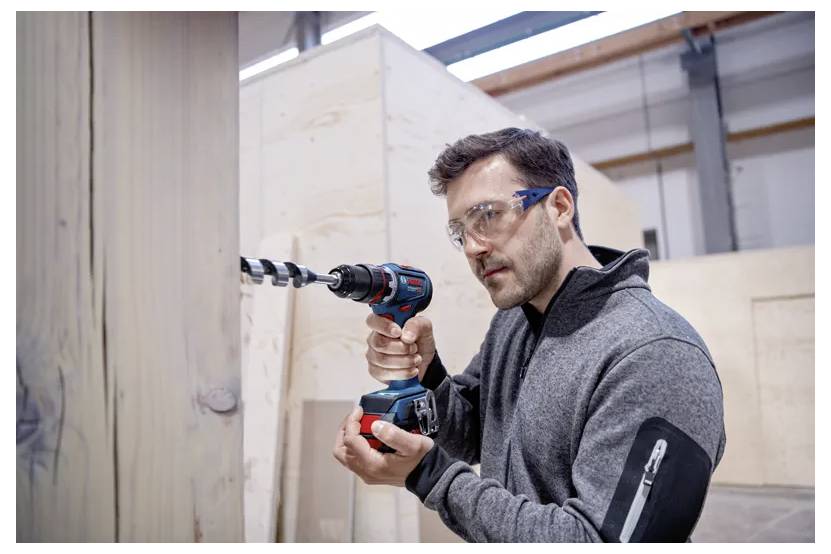 Eine Person mit Sicherheitsbrille benutzt einen Akkuschrauber an einer Holzkonstruktion in einer Werkstatt.