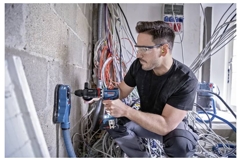 Ein Mann mit Schutzbrille und Gehörschutz bedient ein Elektrowerkzeug zum Bohren in eine Betonwand, umgeben von verschlungenen Kabeln in einer Werkstatt.