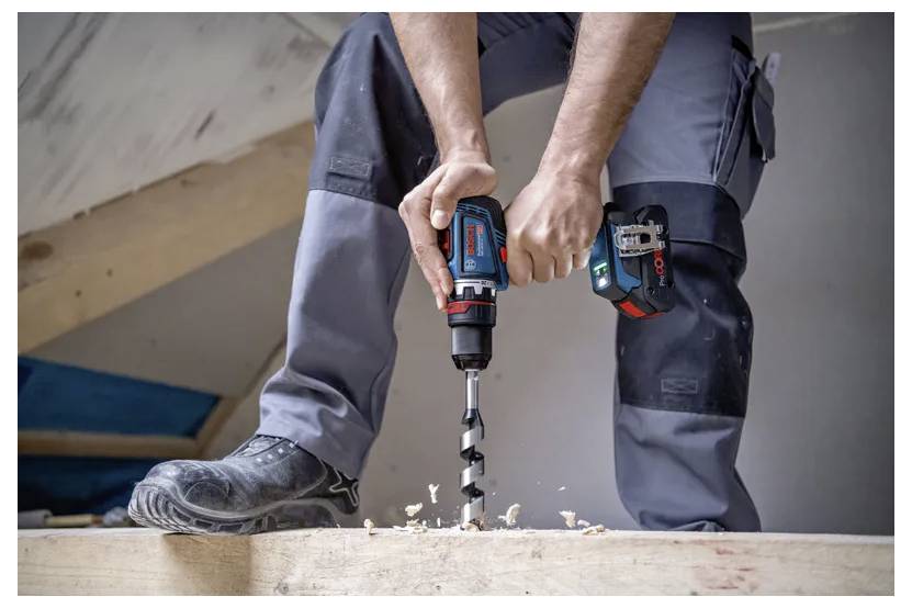 Eine Person in Arbeitshose und Stiefeln verwendet eine Bohrmaschine mit einem großen Bohrer, um in eine Holzplatte zu bohren, während Holzspäne ringsum verstreut sind.