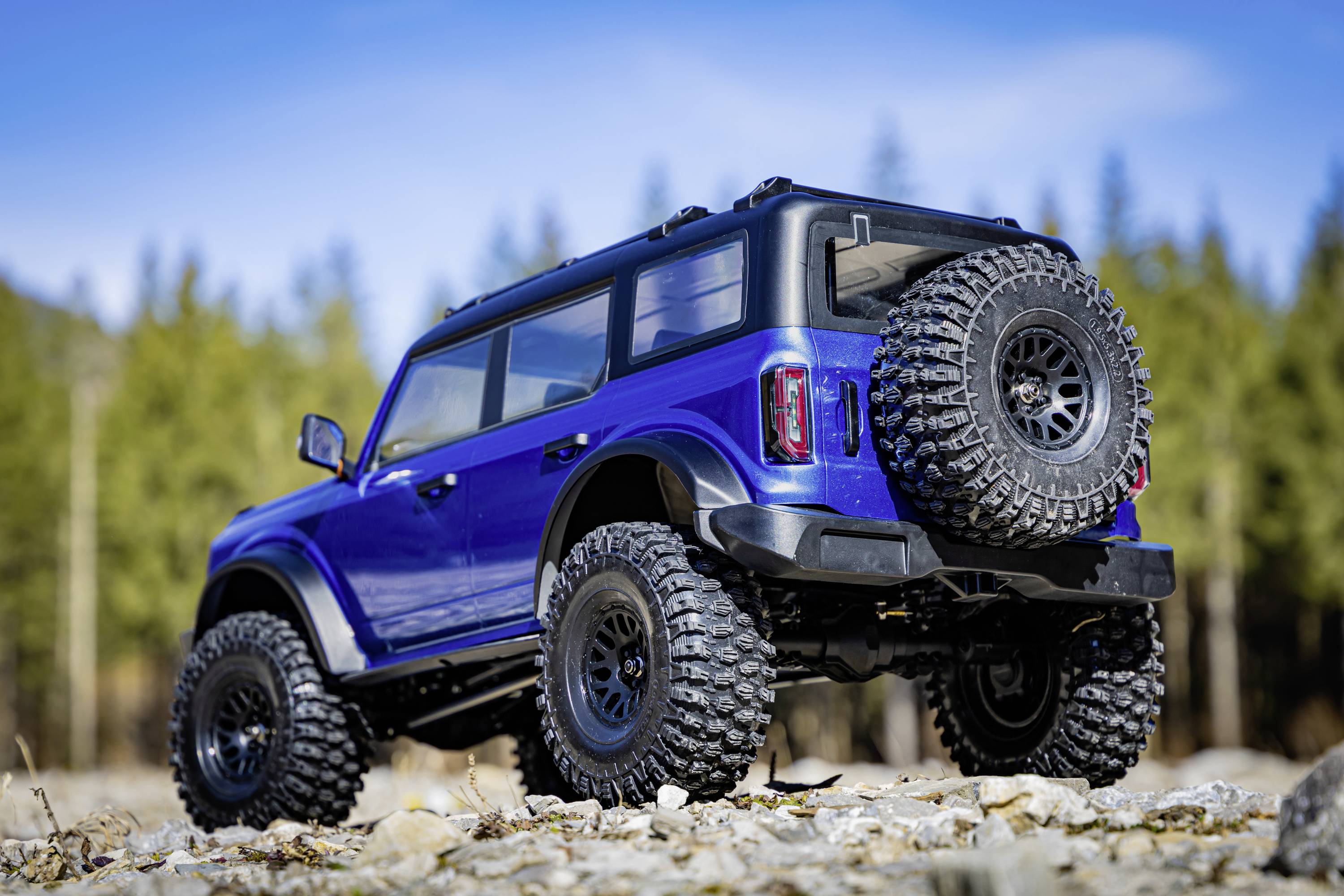 Ein blauer Geländewagen mit großen Reifen steht auf einem kiesigen Weg, umgeben von einem Wald mit Bäumen im Hintergrund unter blauem Himmel.