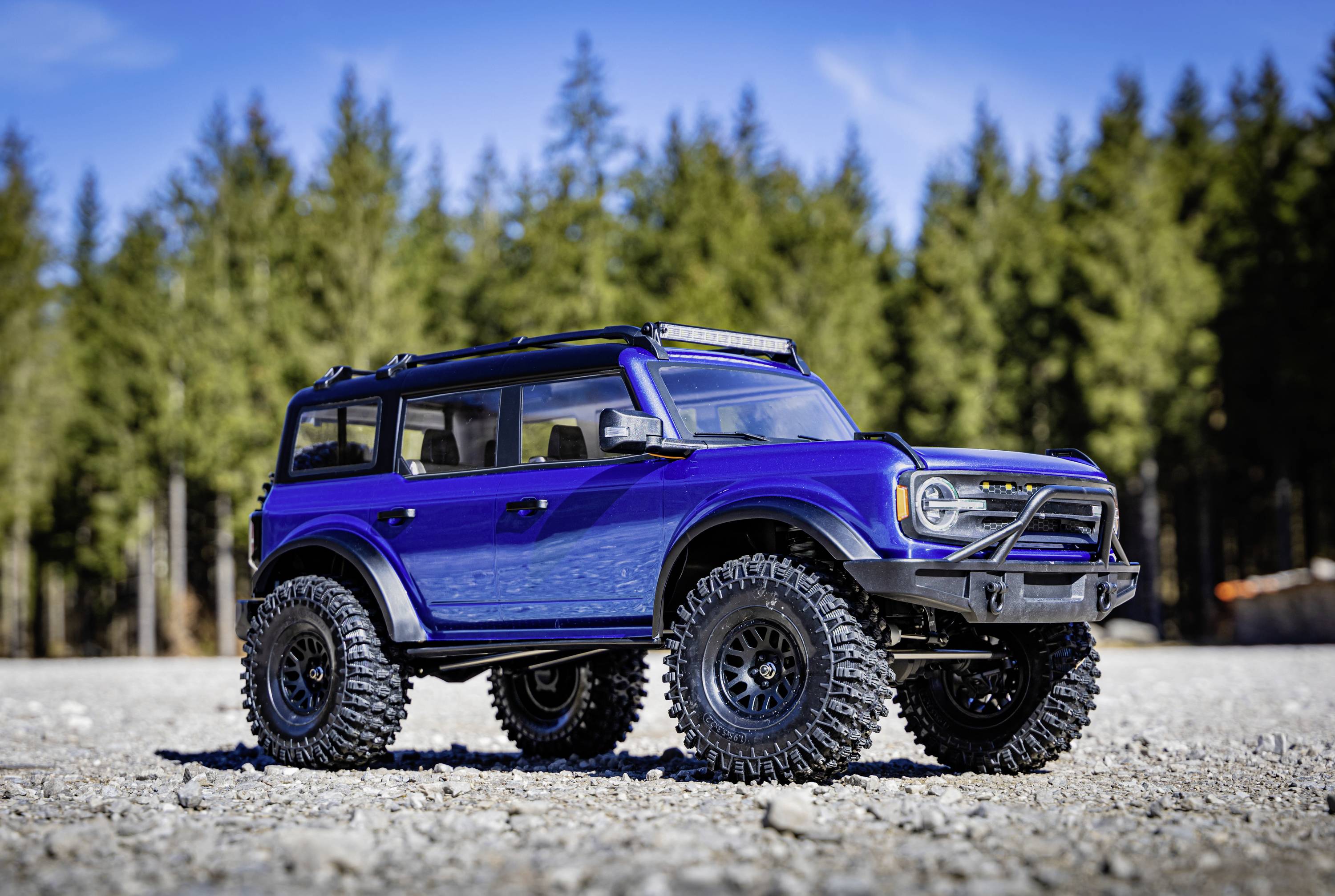 Ein blauer Geländewagen steht auf einem steinigen Boden vor einem Hintergrund aus Nadelbäumen und blauem Himmel.