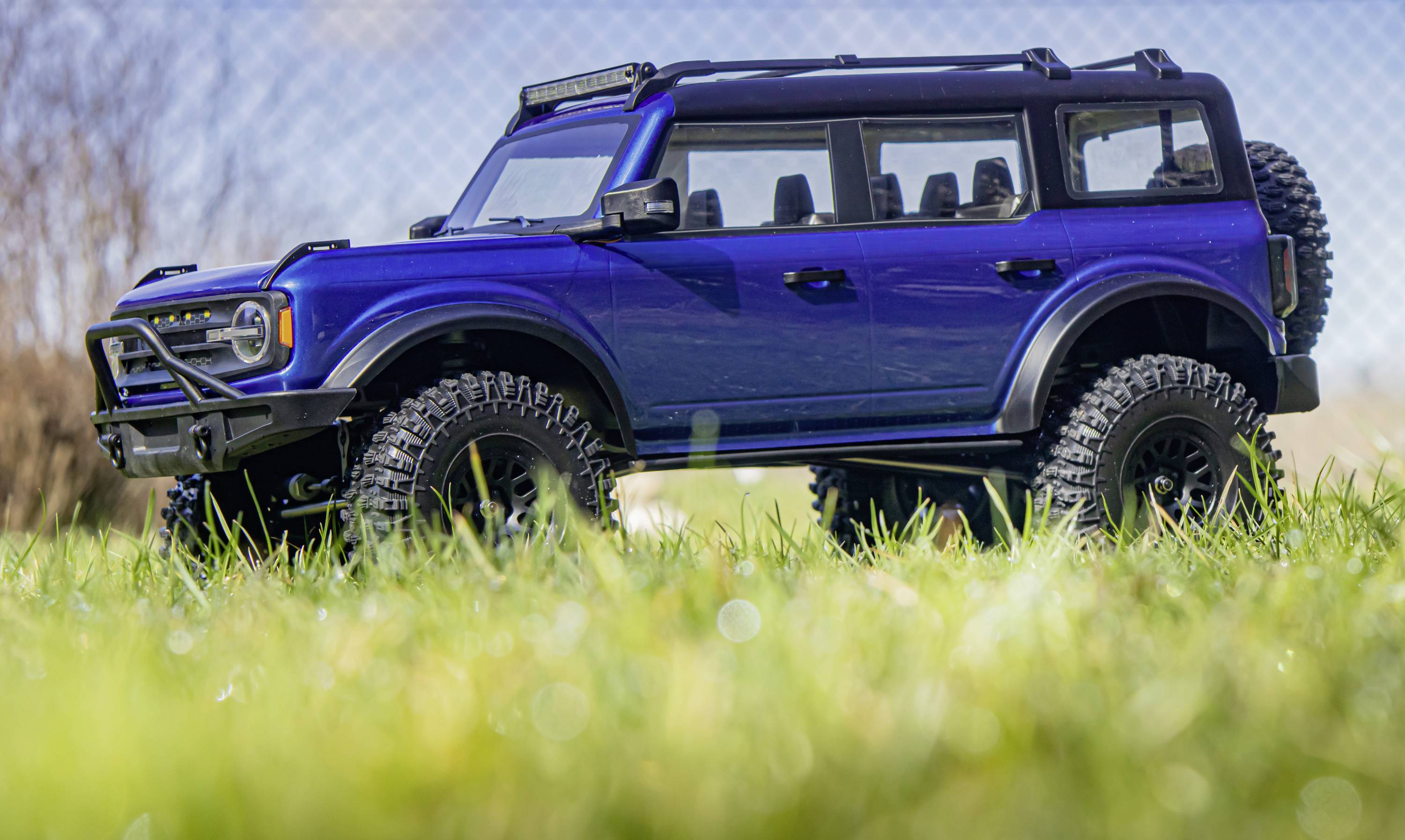 Ein blauer Geländewagen steht auf einer Wiese. Das Fahrzeug hat große Reifen und ist bereit für Offroad-Abenteuer. Im Hintergrund ist ein Drahtzaun zu sehen.