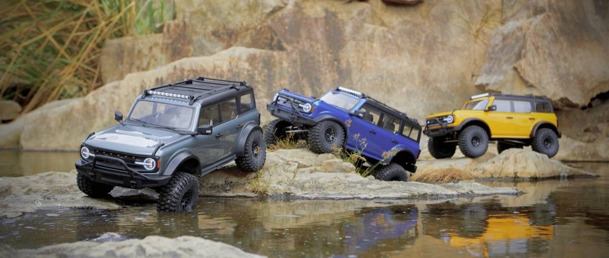 Drei Spielzeug-Geländewagen in unterschiedlichen Farben stehen auf steinigen Felsen an einem kleinen Wasserlauf, hinterlegt von Naturkulisse.