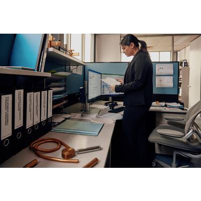 Eine Frau im Büro steht an einem Schreibtisch, blickt auf Dokumente vor zwei Computerbildschirmen. Der Schreibtisch ist unordentlich.