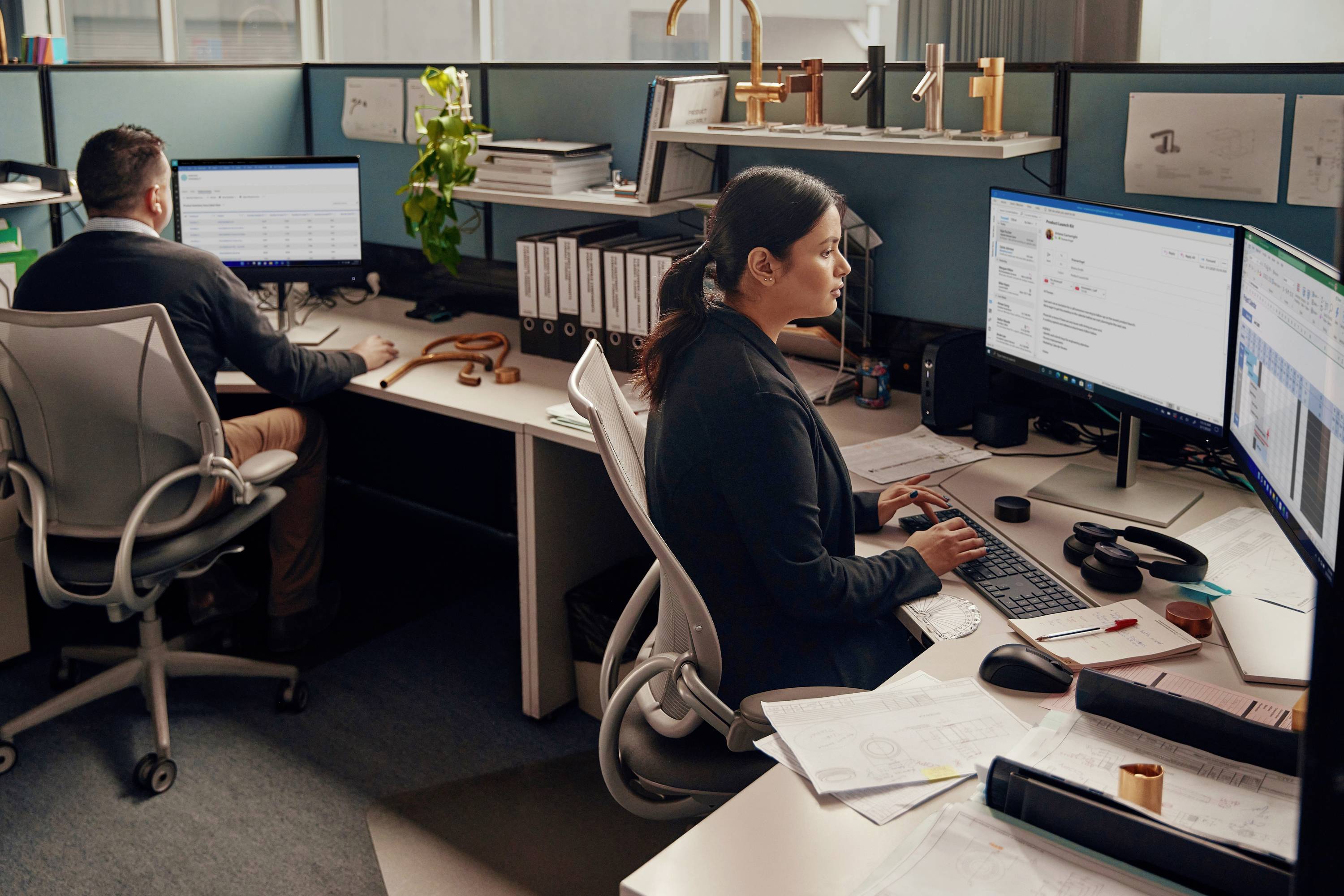 Zwei Personen arbeiten an Computern in einem Büro mit mehreren Monitoren und Schreibtischen voller Unterlagen und Aktenordner.