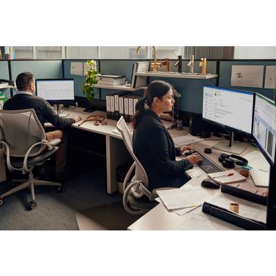 Zwei Personen arbeiten an Computern in einem Büro mit mehreren Monitoren und Schreibtischen voller Unterlagen und Aktenordner.