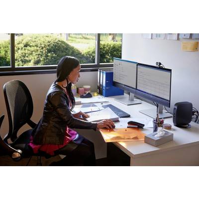 Eine Person arbeitet an einem Schreibtisch mit zwei Monitoren. Büroausstattung und Ordner sind im Hintergrund zu sehen.