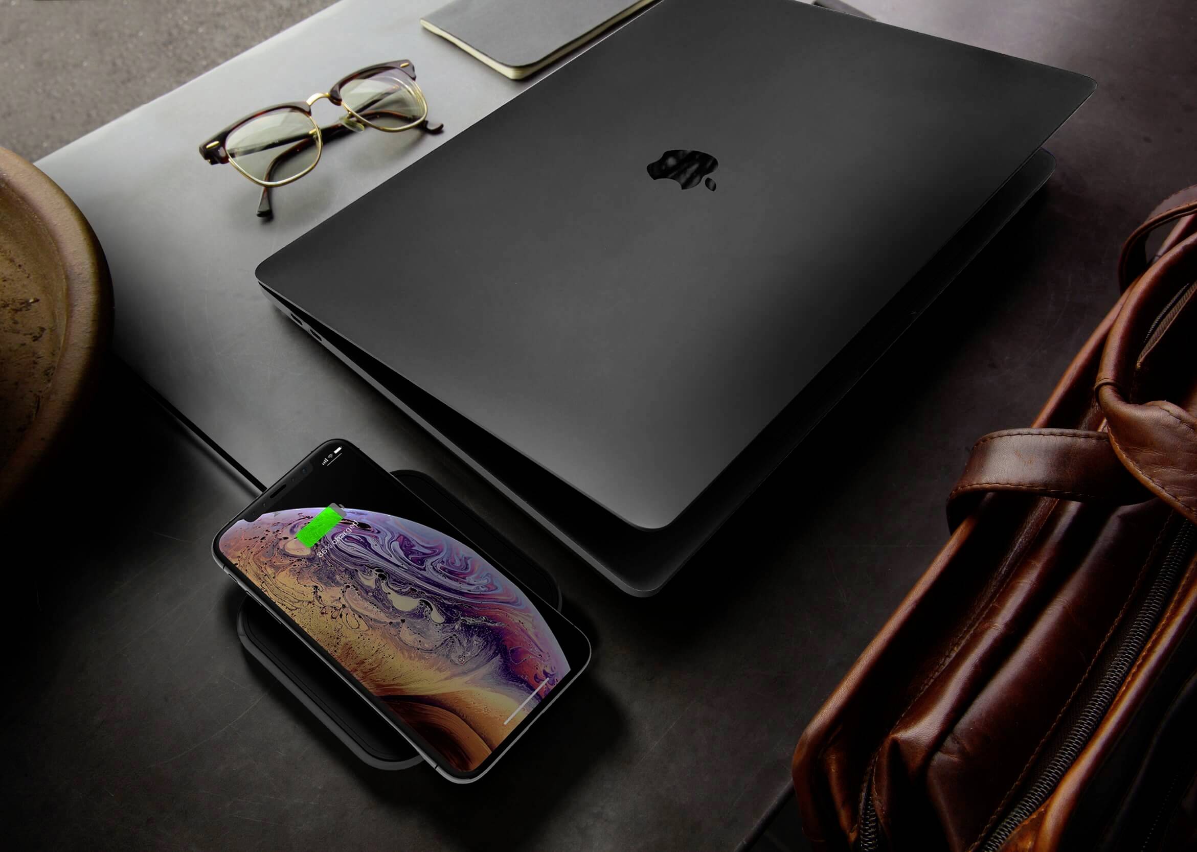 Ein schwarzer Laptop auf einem Tisch neben einer Brille und einem Notizblock. Ein Smartphone lädt drahtlos auf einer Ladestation.
