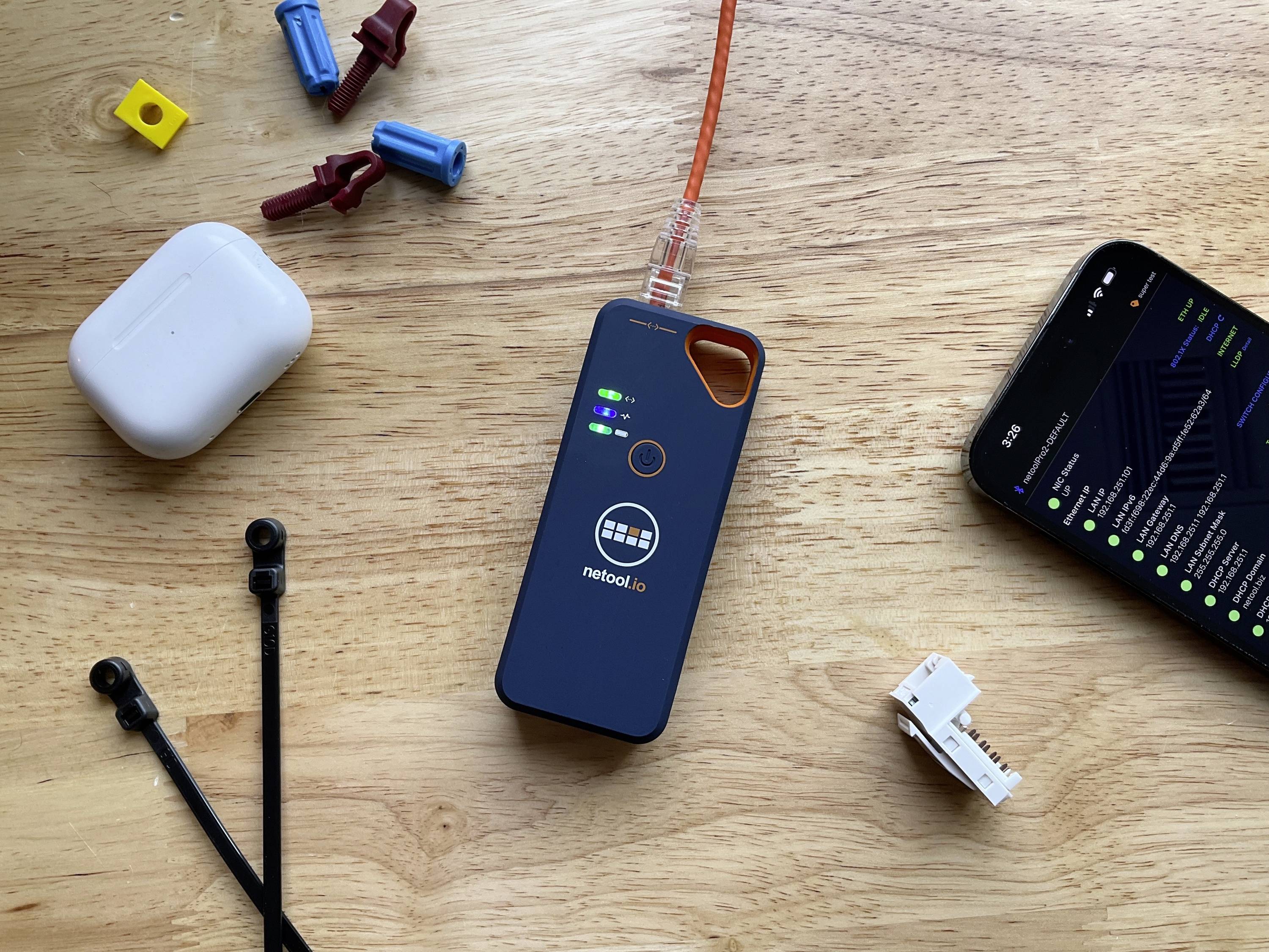 Netzwerktester mit leuchtenden Indikatoren liegt auf Holztisch, umgeben von Kabelbindern, Steckverbindern, einem Smartphone und Kopfhörern.