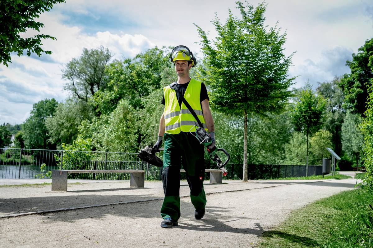 Ein Arbeiter in Sicherheitskleidung und Helm trägt Werkzeuge, während er auf einem Weg in einem grünen Park läuft.