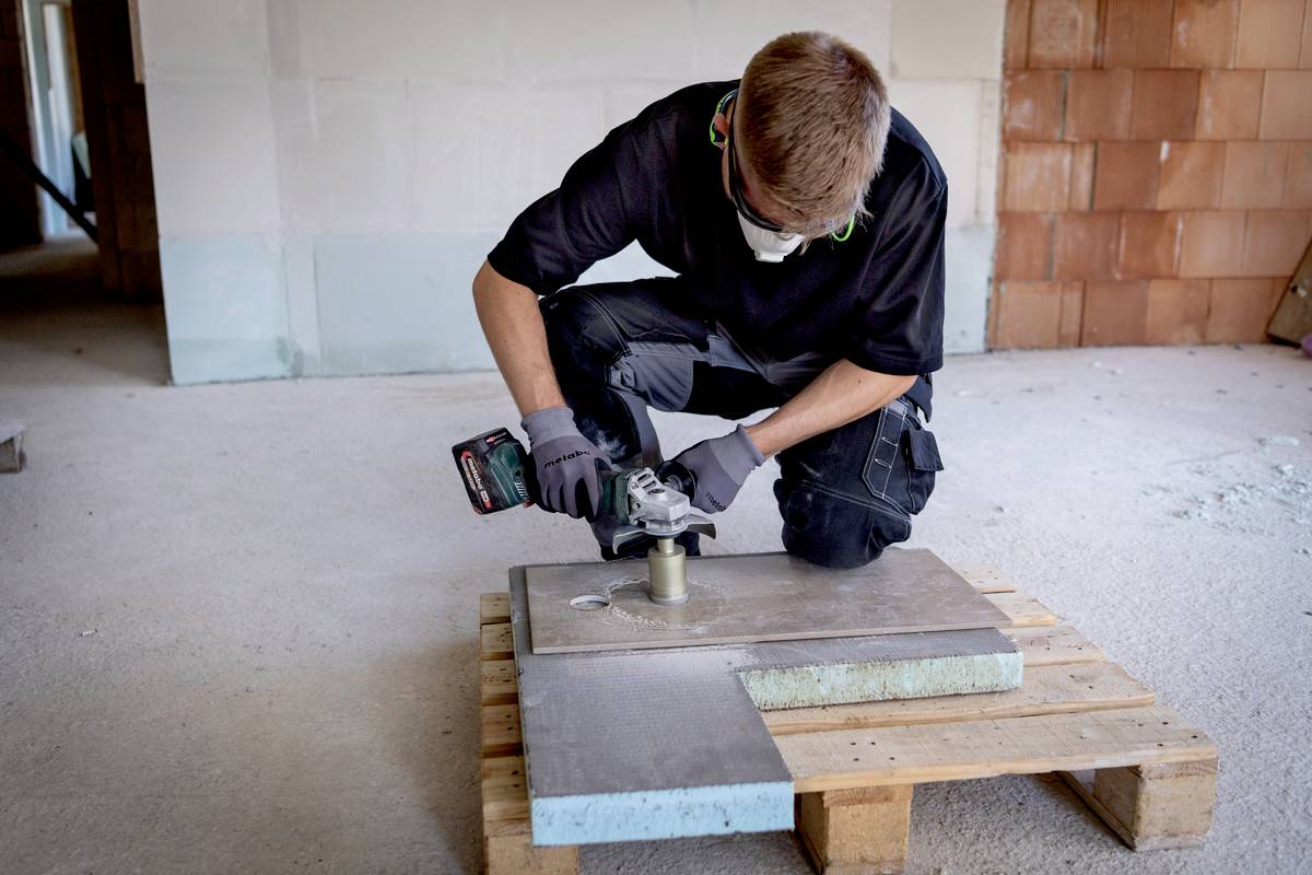 Eine Person mit Schutzmaske bearbeitet mit einer elektrischen Schleifmaschine auf einem Holzpalettentisch ein Metallrohr.
