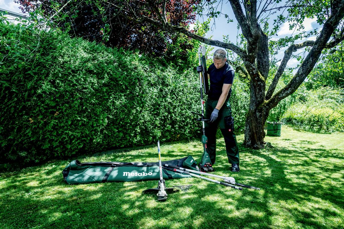 Eine Person im Garten montiert ein Gartenwerkzeug neben einem Baum. Verschiedene Werkzeuge liegen auf dem Gras.