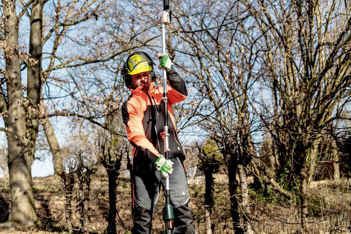 Eine Person mit Schutzkleidung führt Vermessungsarbeiten an einem sonnigen Tag in einem bewaldeten Gebiet durch.
