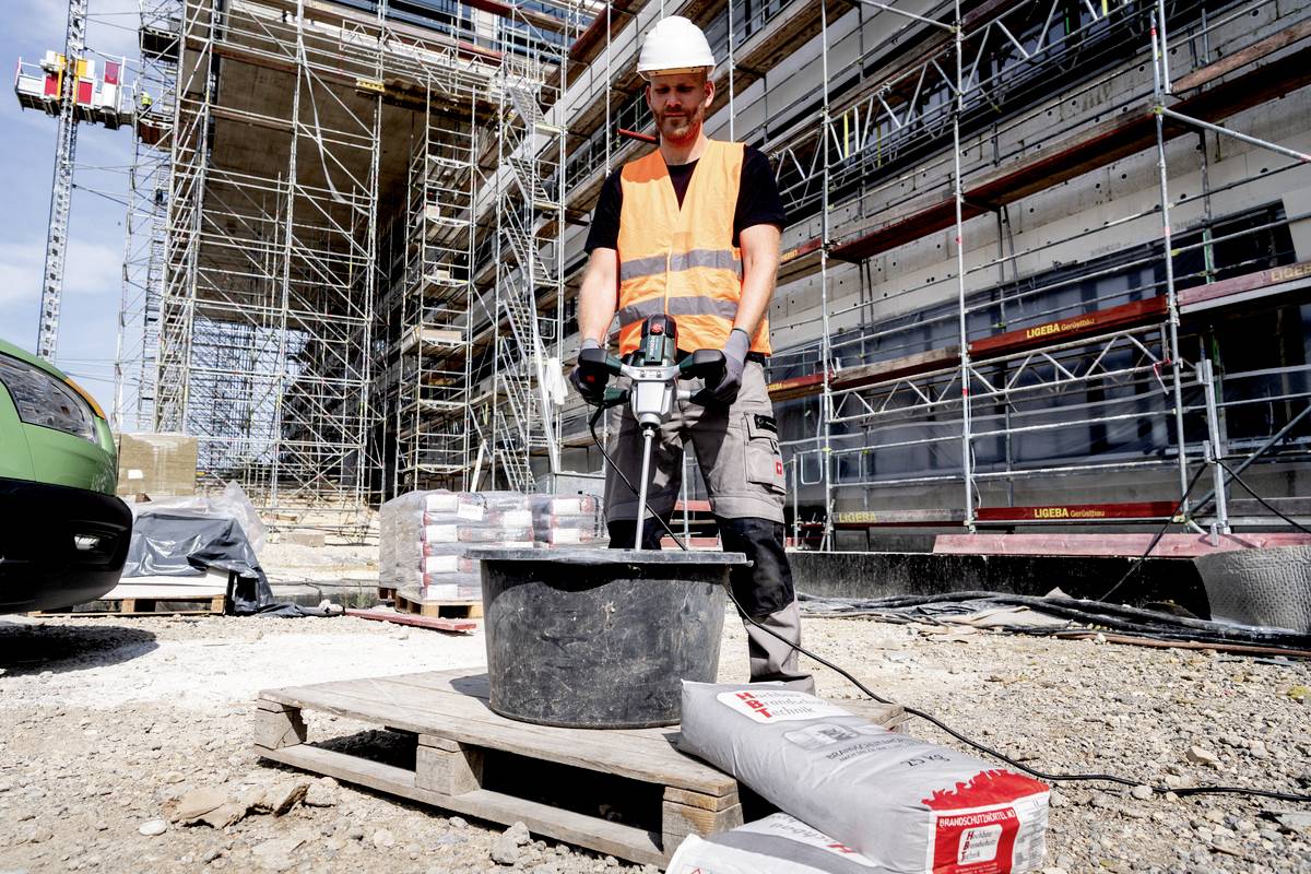 Ein Bauarbeiter mischt Beton auf einer Baustelle neben einem Gerüst und einem im Bau befindlichen Gebäude.