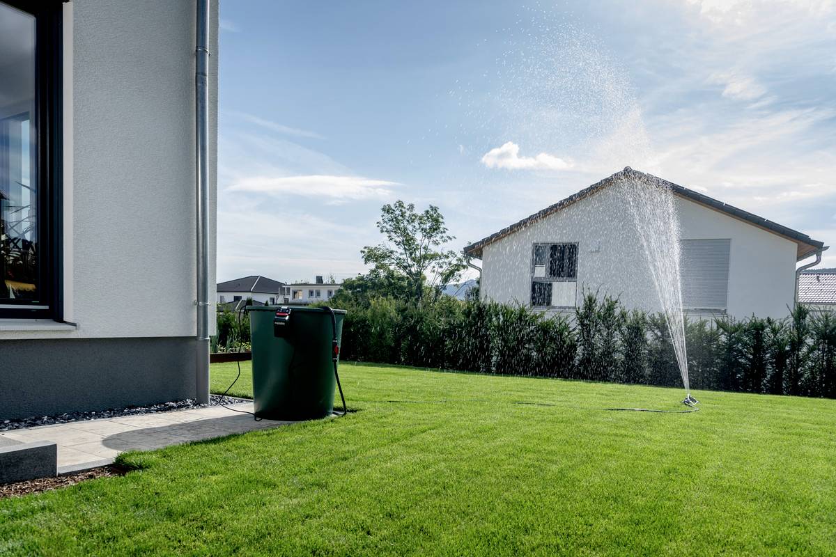 Ein gepflegter Rasen mit einem Rasensprenger, der Wasser sprüht. Rechts im Hintergrund steht ein Wohnhaus, links ein Regentank.