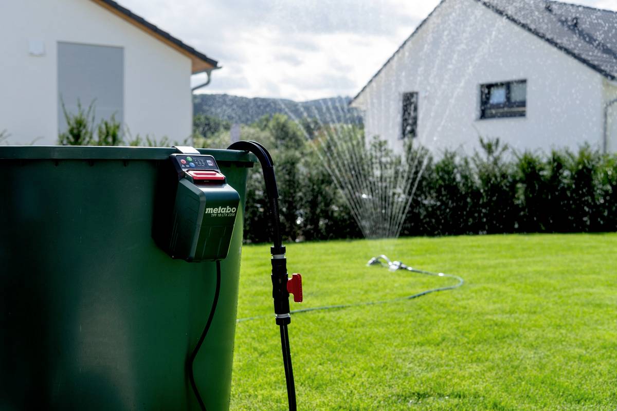 Eine Gartenbewässerungspumpe neben einem Wassertank bewässert mit einem Sprinkler einen grünen Rasen vor modernen Häusern.