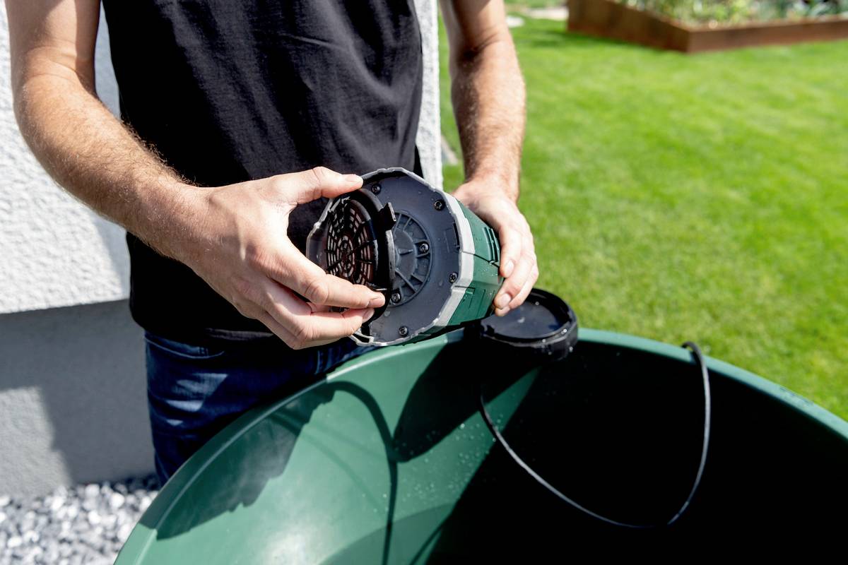 'Person hält Pumpe' über einem grünen Behälter im Garten. Sonne scheint; Fokus liegt auf der Installation oder Wartung einer Wasserpumpe.