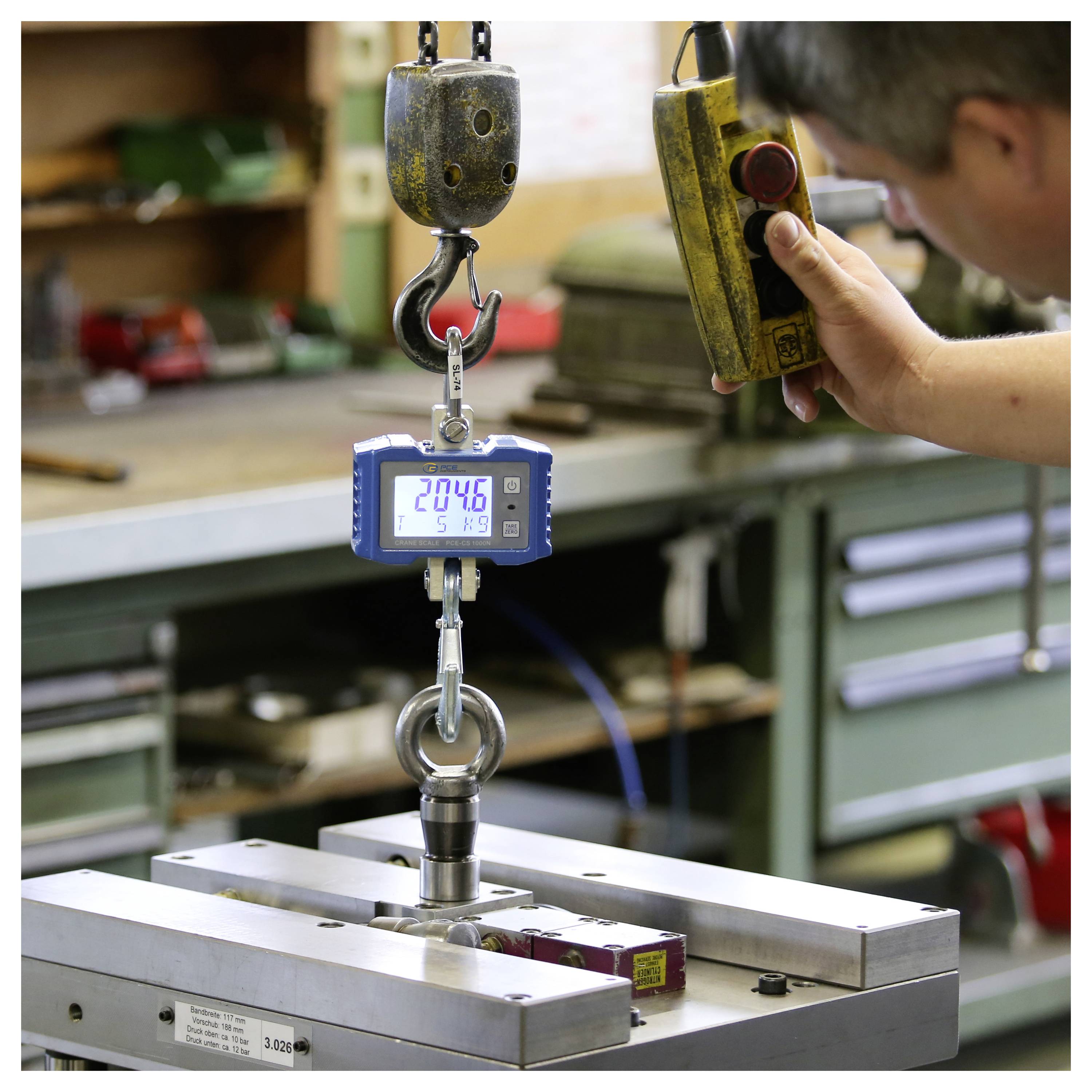 Ein Arbeiter nutzt eine tragbare Fernbedienung, um ein hängendes digitales Kraftmessgerät zu testen, das ein Metallteil in einer Werkstatt hebt.