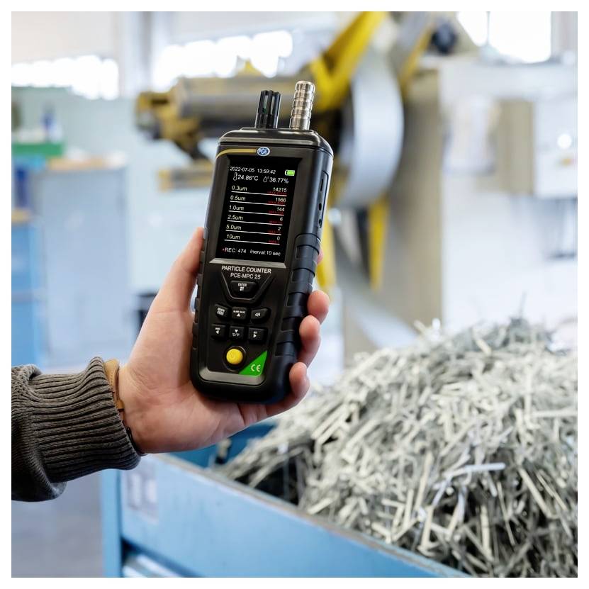 Hand, die einen Partikelzähler in einer Fabrik hält, mit einem Bildschirm, der Messwerte neben einem Behälter mit Metallschrott anzeigt.