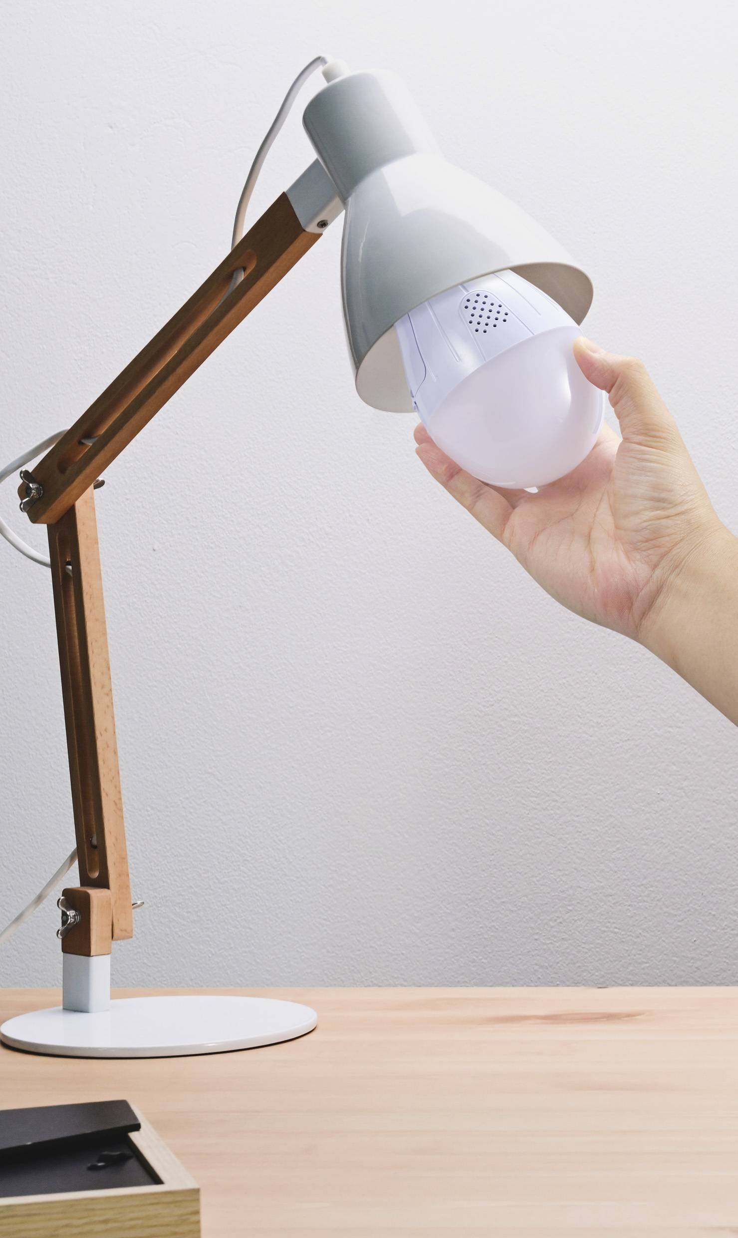 Eine Hand wechselt eine Glühbirne in einer Schreibtischlampe mit Holz- und Metallgestaltung.