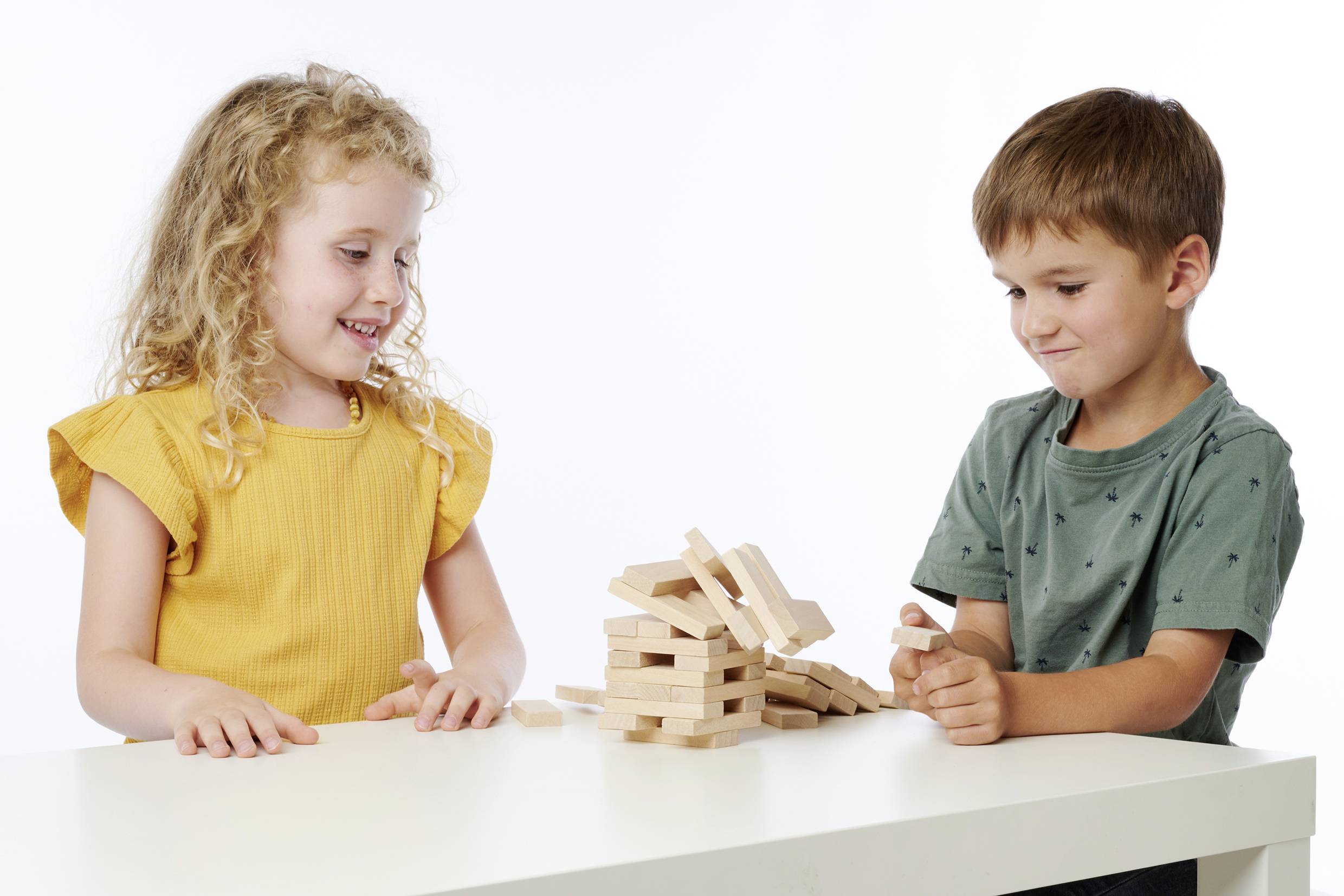 Zwei Kinder spielen mit Holzbausteinen an einem Tisch. Die Bausteine sind teilweise umgefallen. Sie wirken konzentriert und fröhlich.