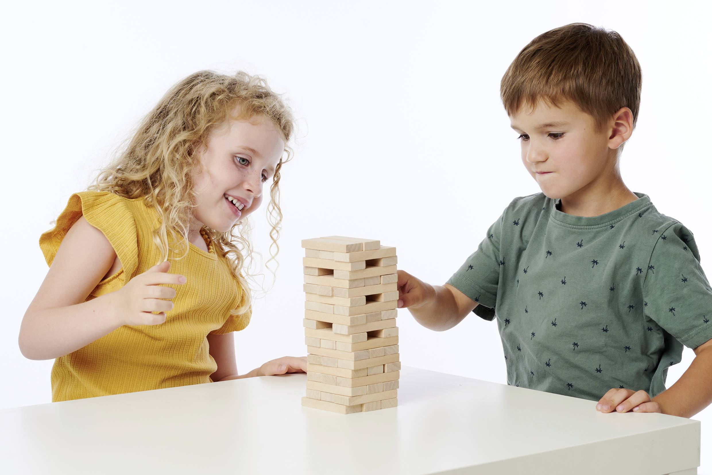 Zwei Kinder spielen ein Tischspiel mit einem Turm aus Holzblöcken. Ein Kind zieht vorsichtig einen Block heraus, während das andere zusieht.