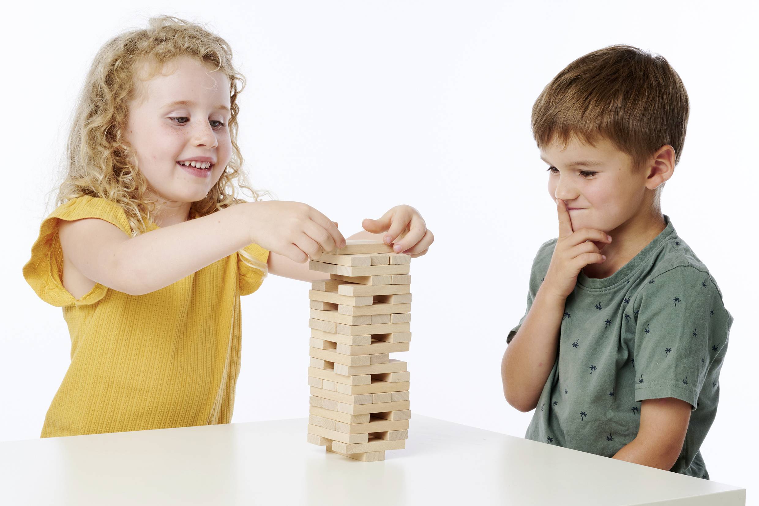 Zwei Kinder spielen Jenga am Tisch. Das Mädchen legt konzentriert einen Holzstein auf den Turm, während der Junge neugierig zusieht.